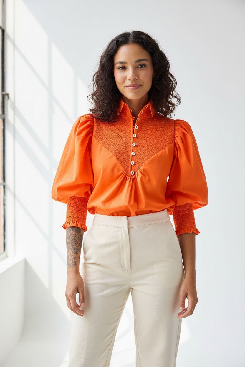 A person with curly hair is seen from behind wearing the HOPE FOR FLOWERS - SMOCKED SHIRT IN FLAME by HOPE FOR FLOWERS—an orange organic cotton poplin shirt with puff sleeves—and light pants. A numbered tattoo is visible on their neck against a plain background.