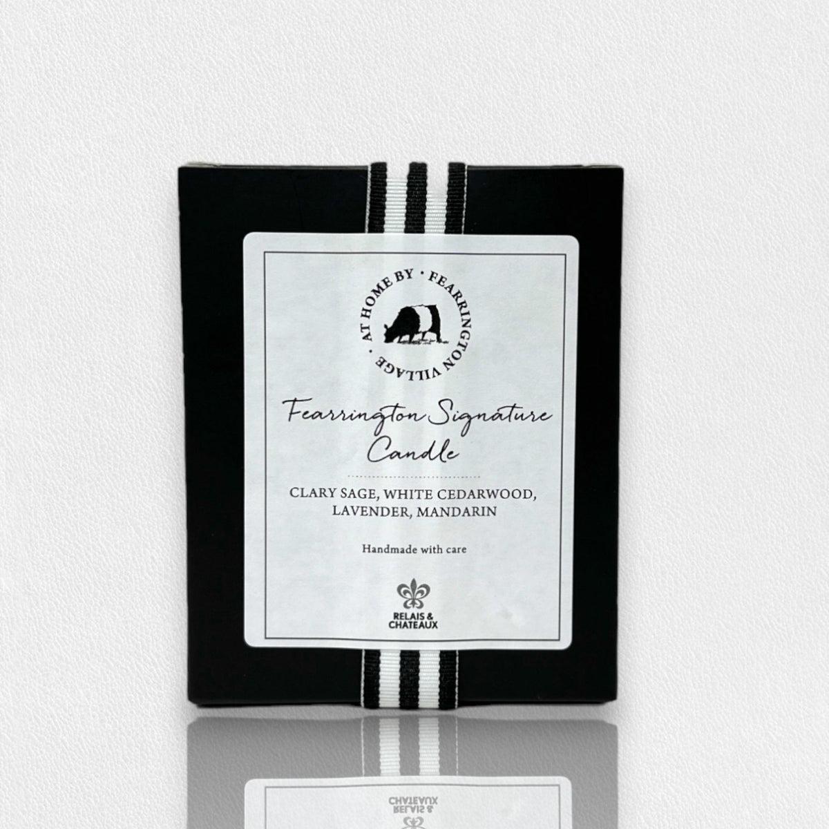 A rectangular black box prominently displays the label &quot;Fearrington Signature Candle&quot; at its center, reading &quot;Clary Sage, White Cedarwood, Lavender, Mandarin.&quot; The top features a white circular logo with the subtitle &quot;Handmade with care.&quot; As part of our luxurious natural soy candles collection, it is crafted by Fearrington Village - Home Decor and includes two vertical white stripes.