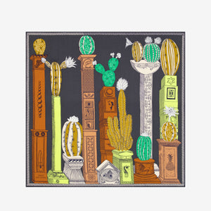 The INOUI EDITIONS - TEQUILA SCARF IN LEMON by INOUI features vibrant cacti and succulents in green, yellow, and white on brown and gray pedestals, set against a dark gray background with a decorative border.