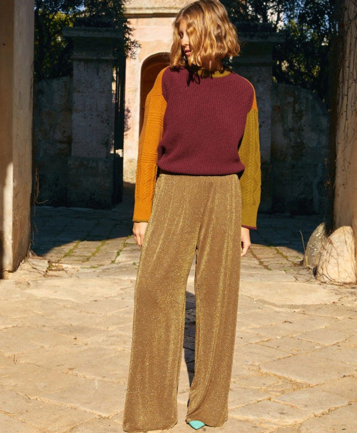 A woman with long brown hair stands against a neutral background, wearing a black ensemble from Momoni that exudes effortless elegance. She sports the comfortable Momoni Lanzarote Pant in Lurex Jersey and an equally elegant lurex jersey top. A patterned scarf is draped around her neck, and she has her hands in her pockets. The outfit, made in Italy, embodies chic sophistication.