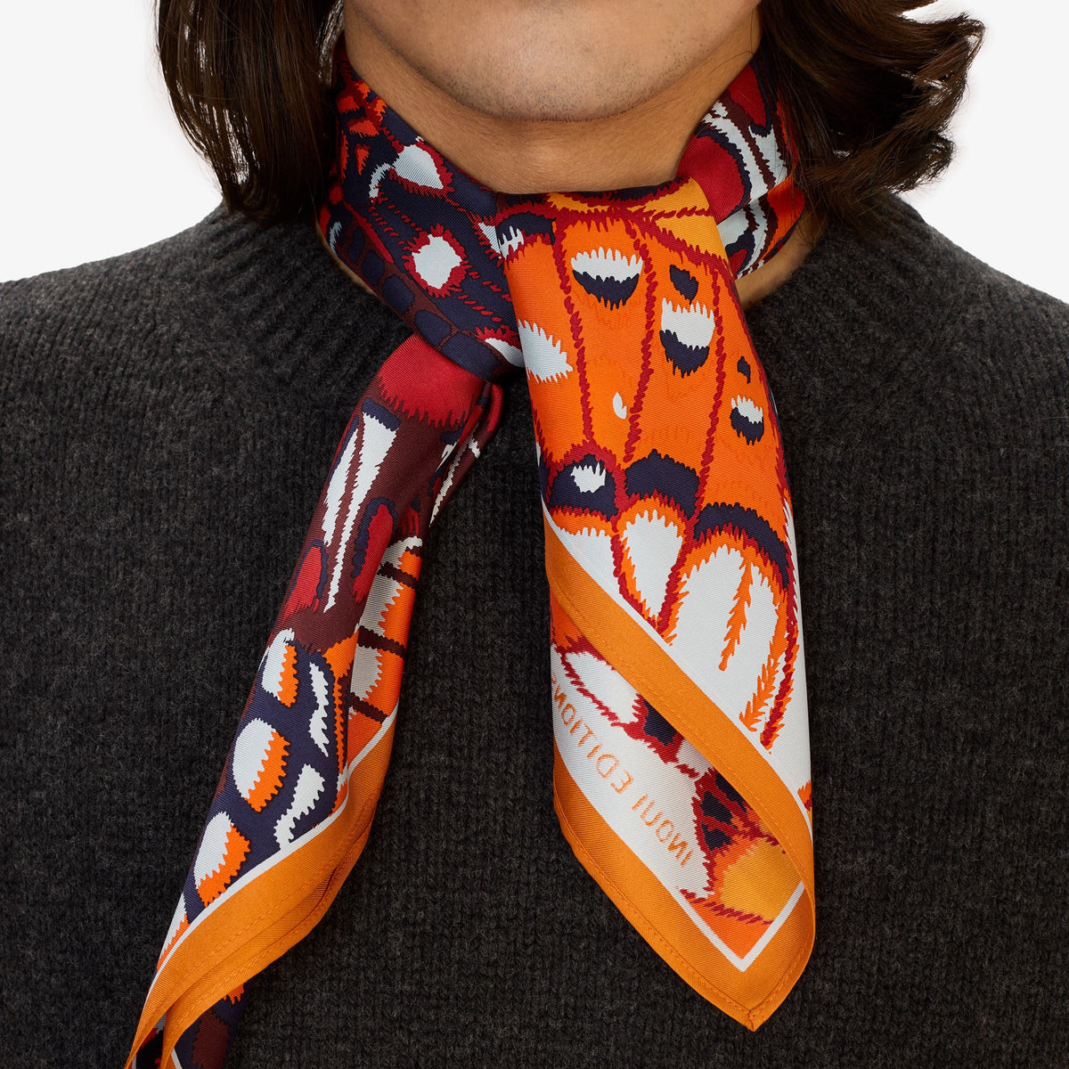 A person in a dark grey knit sweater wears an INOUI EDITIONS PAPILLONS Orange Silk Scarf, featuring bold abstract butterfly designs and “INOUI EDITIONS” on the border, neatly tied around their neck.