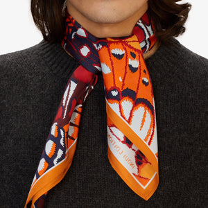 A person in a dark grey knit sweater wears an INOUI EDITIONS PAPILLONS Orange Silk Scarf, featuring bold abstract butterfly designs and “INOUI EDITIONS” on the border, neatly tied around their neck.