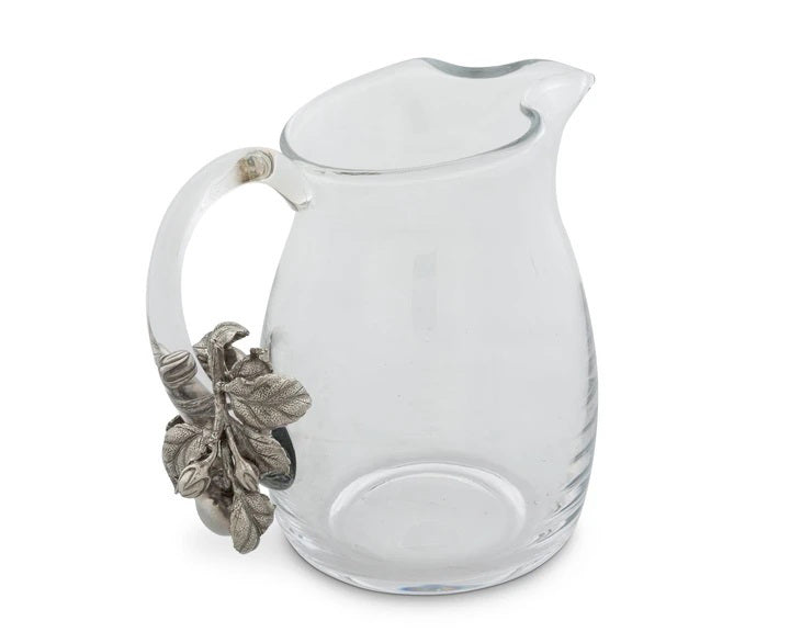The VAGABOND HOUSE - APPLE GLASS PITCHER from VAGABOND HOUSE features a clear glass body with a curved spout and an ornate handle decorated with silver-toned leaves and acorns, blending traditional craftsmanship into a unique, elegant piece of functional tableware.