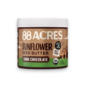 A slice of seeded bread on a wooden board is topped with a creamy vegan spread and three strawberry slices. Nearby are a knife with spread, more bread slices, whole strawberries, and a jar of 88 ACRES Dark Chocolate Sunflower Seed Butter.