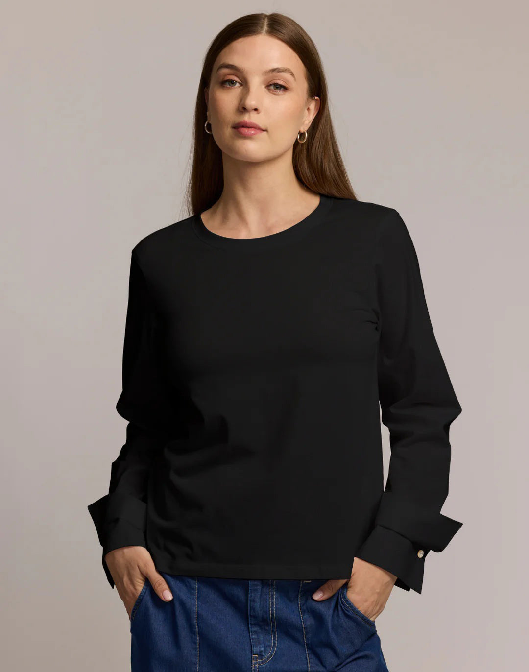 A woman with long brown hair wears the HINSON WU REAGAN LONG SLEEVE FOUNDATION LAYER and blue jeans, standing against a plain light background and looking directly at the camera with a neutral expression.
