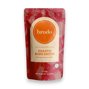 A packet of BRODO BROTH COMPANY’s BRODO BROTH- SIGNATURE HEARTH BONE BROTH sits on a light surface, surrounded by a bowl of chopped vegetables, a cooked turkey leg, half an onion, a bay leaf, and a cup of rich bone broth made from grass-fed beef and organic chicken.