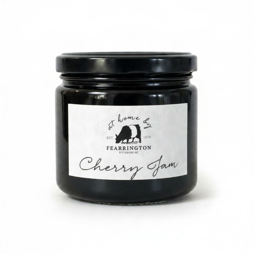 A glass jar with a black lid labeled “FEARRINGTON SIGNATURE COLLECTION - FEARRINGTON HOUSE CHERRY JAM” from Fearrington House, featuring a cow silhouette and the phrase “at home by Fearrington, Pittsboro, NC, EST. 1974,” on a white background. This versatile spread is crafted from ripe cherries to bring a deliciously sweet flavor to your pantry.