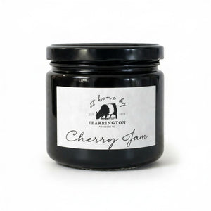 A glass jar with a black lid labeled “FEARRINGTON SIGNATURE COLLECTION - FEARRINGTON HOUSE CHERRY JAM” from Fearrington House, featuring a cow silhouette and the phrase “at home by Fearrington, Pittsboro, NC, EST. 1974,” on a white background. This versatile spread is crafted from ripe cherries to bring a deliciously sweet flavor to your pantry.