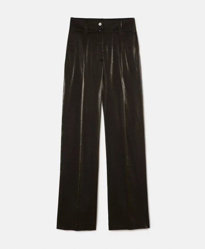 A person stands against a light-colored stone wall, wearing a loose-fitting, long-sleeved brown top with bio-galalite buttons paired with the high-waisted, wide-leg MANHATTAN PANT from MOMONI. They have long hair and are looking directly at the camera. The background is simple and uncluttered.