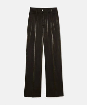 A person stands against a light-colored stone wall, wearing a loose-fitting, long-sleeved brown top with bio-galalite buttons paired with the high-waisted, wide-leg MANHATTAN PANT from MOMONI. They have long hair and are looking directly at the camera. The background is simple and uncluttered.