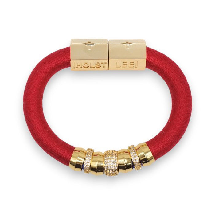 The HOLST + LEE - LUXE CLASSIC BRACELET by HOLST &amp; LEE features an elegant orange cord with gold-plated accents, including a rectangular gold clasp adorned with several gold and diamond-encrusted beads near the clasp.