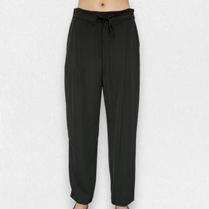 A person is wearing the MEIMEIJ - DRAWSTRING TROUSER PANT from the brand MEIMEIJ. These black, high-waisted trousers feature a loose fit and are made from a blend of polyester and elastane, complete with a tie belt. The image is taken from the waist down, showcasing the trousers in full against a plain white background, with the person's feet partially visible at the bottom.