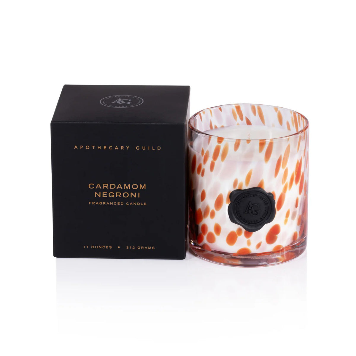 A ZODAX APOTHECARY GUILD Opal Glass Small Candle in Cardamom Negroni with orange and white spots sits beside its black box. Enjoy this beautifully scented candle’s long burn time and elegant design.