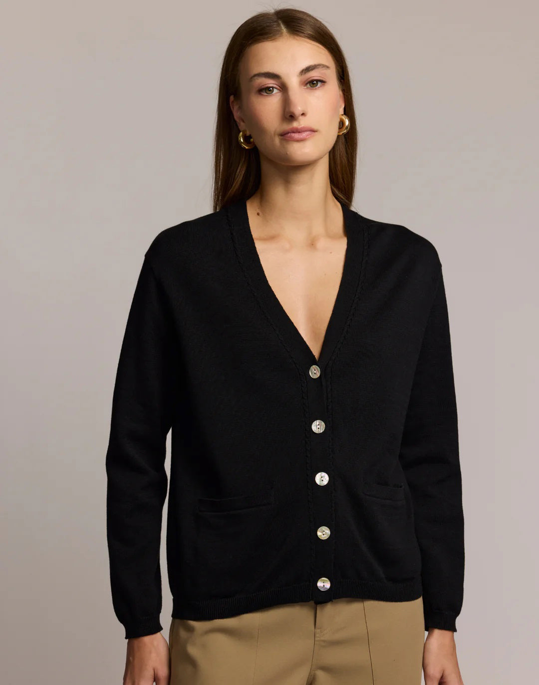 A woman with straight brown hair wears the HINSON WU CASSIE Long Sleeve Cotton Silk Cardigan in camel, styled with black pants. She poses confidently, one hand to her chin, against a neutral backdrop.