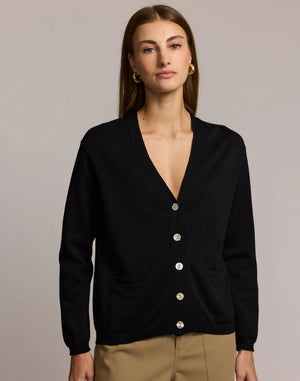 A woman with straight brown hair wears the HINSON WU CASSIE Long Sleeve Cotton Silk Cardigan in camel, styled with black pants. She poses confidently, one hand to her chin, against a neutral backdrop.