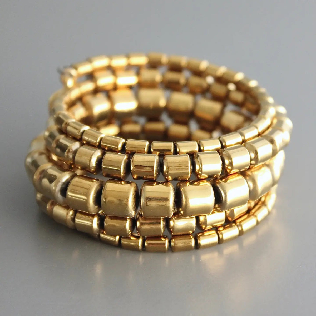 The DAVID AUBREY Gold Plated Hematite Wrap Bracelet features shiny gold beads in four stacked rows, creating a chunky, textured look. This handcrafted piece from New Jersey is shown against a plain gray background.