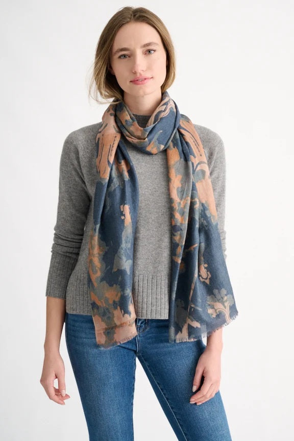 A woman stands against a plain white background, wearing a gray sweater, blue jeans, and the KINROSS Cashmere Tapestry Scarf in Navy by KINROSS.