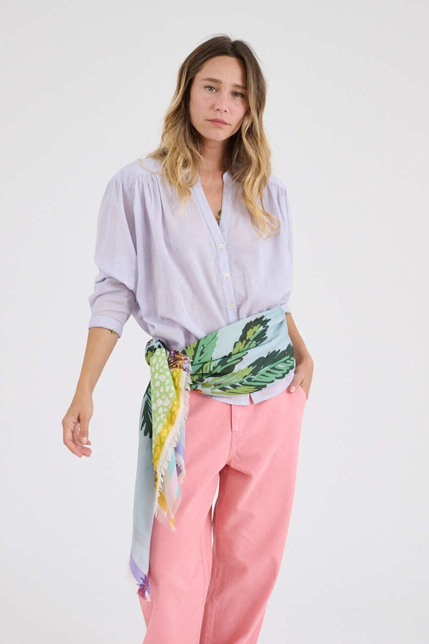 A woman with light brown hair wears a pale purple button-up shirt, pink pants, and the STORIATIPIC - CALI MAX SCARF—a large, colorful tropical-print accessory by STORIATIPIC—draped around her shoulders against a plain white background.