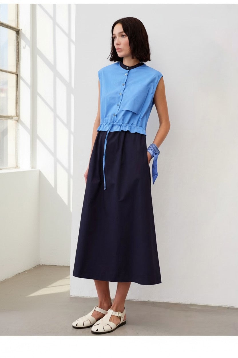 A woman stands indoors by a window wearing the FLOOR - COLOR BLOCK COTTON DRESS IN BLUE from FLOOR. Light streams through the window onto the floor.