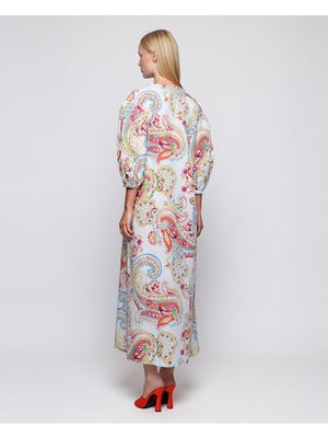 A woman faces forward in the MIRTO 1956 Paisley Print Long Sleeve Dress, featuring multicolor linen, puffed three-quarter sleeves, and a V neckline. She has straight blonde hair, red shoes, and stands against a plain white background.