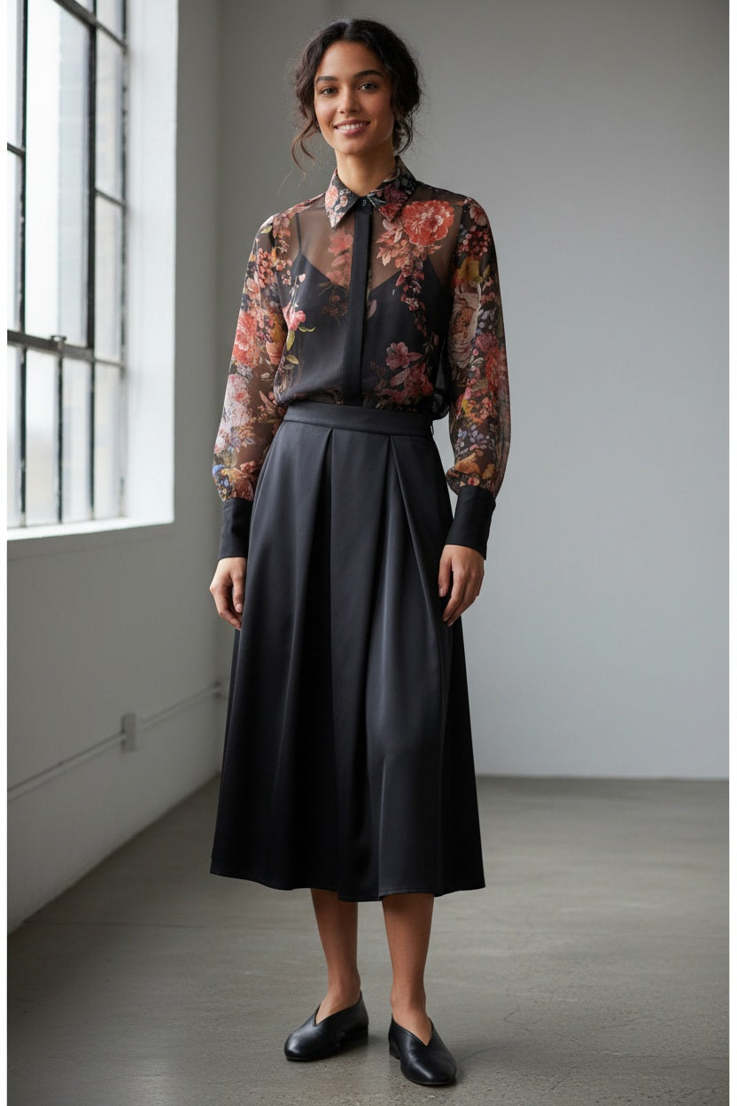 A woman with wavy dark hair models the MILVA MI - FLORAL VOILE BLOUSE by MILVA—a semi-sheer, long-sleeve top with a dark floral print and black collar—paired with a high-waisted skirt, standing before a plain gray background.
