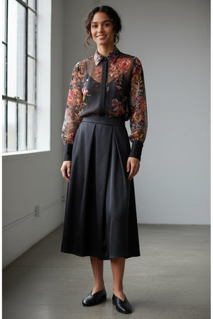 A woman with wavy dark hair models the MILVA MI - FLORAL VOILE BLOUSE by MILVA—a semi-sheer, long-sleeve top with a dark floral print and black collar—paired with a high-waisted skirt, standing before a plain gray background.