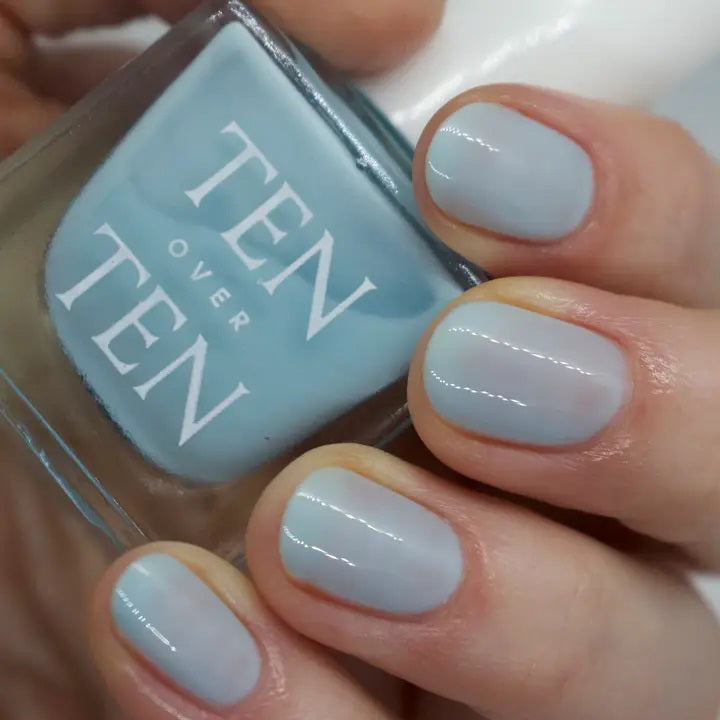 A glass bottle of TENOVERTEN - NAIL POLISH from the brand TEN OVER TEN, featuring a white cap and a dusty pink nail color. The front label displays "TEN OVER TEN" in white uppercase letters. This vegan, eight-free formula ensures both beauty and peace of mind. The background is plain and light-colored.