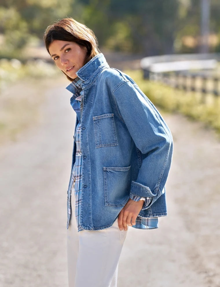 A woman stands indoors wearing the FRANK & EILEEN Bantry Barn Jacket in California Vintage Denim 1978 Wash over a white top, blue jeans, and brown ankle boots. She has short, dark hair and is smiling as natural light streams through a large window beside her.