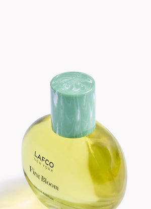 A box of LAFCO - FINE FRAGRANCE, FIRST BLOOM 50ML stands among white flowers and green vines against a pale beige background.