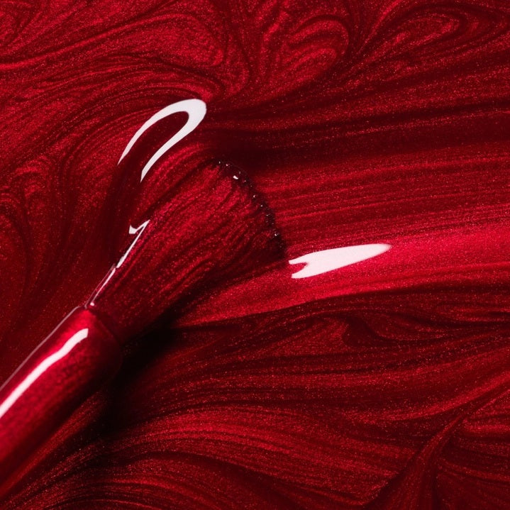 A hand with shiny, iridescent red nails holds a matching bottle labeled “MANUCURIST - GREEN™ NAIL POLISH IN RED HIBISCUS” by MANUCURIST INC against brown skin. This luxurious vegan, cruelty-free nail polish delivers vibrant color and a flawless finish.