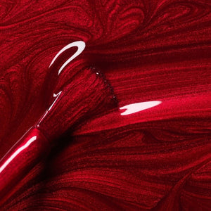 A hand with shiny, iridescent red nails holds a matching bottle labeled “MANUCURIST - GREEN™ NAIL POLISH IN RED HIBISCUS” by MANUCURIST INC against brown skin. This luxurious vegan, cruelty-free nail polish delivers vibrant color and a flawless finish.
