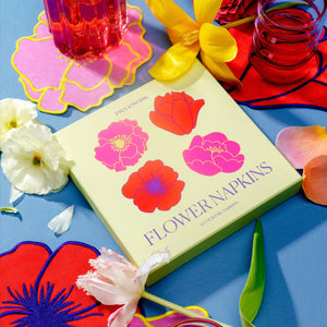 Colorful floral coasters sit on a blue surface with glasses of red liquid, surrounded by scattered white tulips and PIECEWORK PUZZLES' LINEN FLOWER COCKTAIL NAPKINS for a vibrant tabletop display.