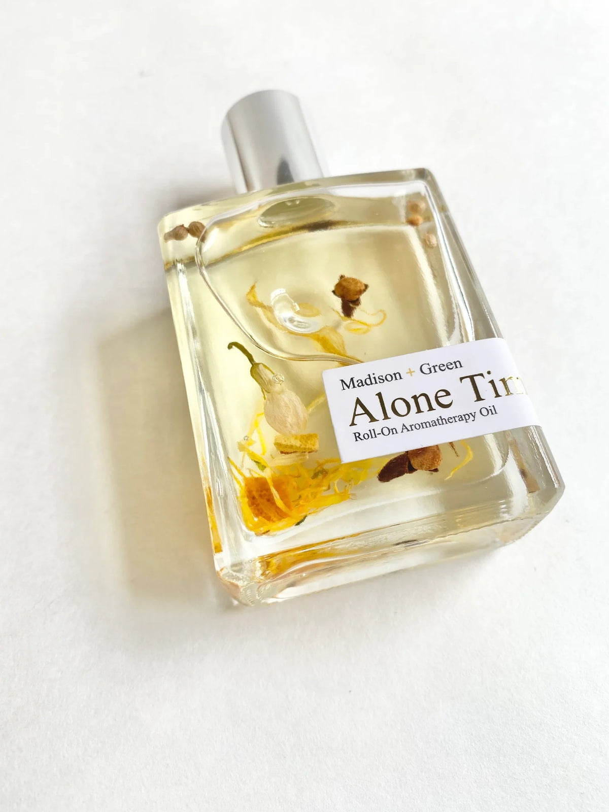 A clear glass bottle with a silver cap holds yellow relaxing body oil infused with small flower petals. The label reads “MADISON AND GREEN - ALONE TIME RELAXING AROMATHERAPY BODY OIL MINI,” highlighting an essential oil blend by MADISON GREEN crafted to help you unwind and relax.