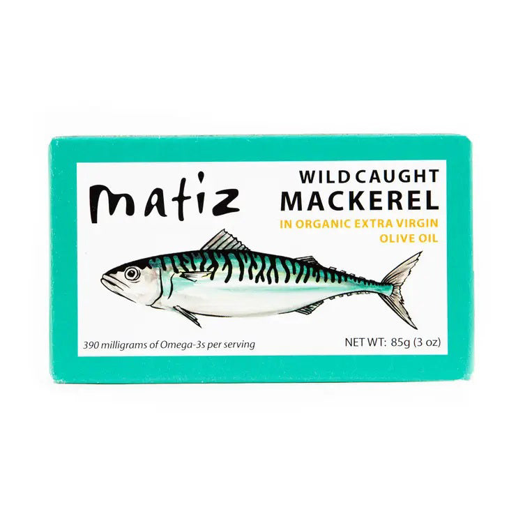 The opened tin of MATIZ - Wild Caught Mackerel in Organic Olive Oil reveals tender fish slices, with an unopened package featuring a mackerel illustration beside it. A fork, crusty bread, and lemon slices are elegantly arranged on a pristine white background. Made in Spain by MATIZ ESPANA with care.