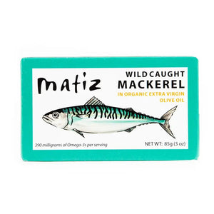 The opened tin of MATIZ - Wild Caught Mackerel in Organic Olive Oil reveals tender fish slices, with an unopened package featuring a mackerel illustration beside it. A fork, crusty bread, and lemon slices are elegantly arranged on a pristine white background. Made in Spain by MATIZ ESPANA with care.
