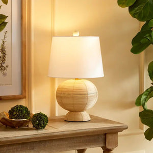 The NAPA HOME AND GARDEN Maye Rattan Hourglass Lamp with Shade features a wicker-style base and white shade, casting a warm glow on any wooden surface.