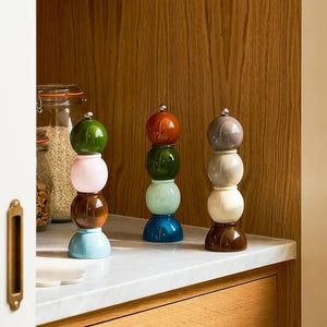 Three vibrant ADDISON ROSS LONDON Bobbin Salt or Pepper Mills (24cm) from ADDISON ROSS are stacked on a kitchen counter near wooden cabinets, with jars of dry food visible in the background.