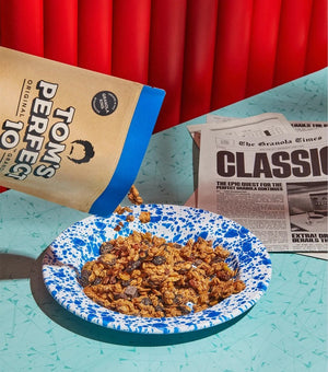 A beige and blue resealable pouch labeled "TOM'S PERFECT 10 - CLASSIC GRANOLA," featuring a transparent window at the bottom displaying the granola inside mixed with cherries. The pouch weighs 10 ounces (283.5 grams). A circular label says "Original Formula.