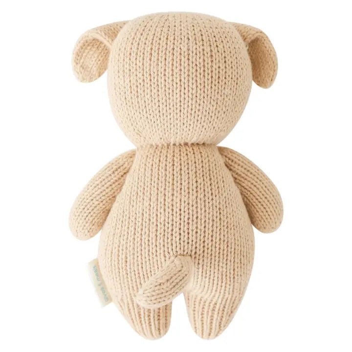 The CUDDLE + KIND - HANDMADE BABY PUPPY by CUDDLE &amp; KIND is displayed from the back, highlighting its beige knitted body, small floppy ears, stubby arms, and short tail with no visible facial details. Meticulously hand-knit from 100% cotton yarn, its texture is distinctly visible, enhancing its cozy and handcrafted appeal. Perfect for those who appreciate fair trade and hand-knit baby animals.