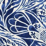 A person with wavy blonde hair stands outdoors, dressed in a sleeveless SILK PRINTED BLOUSE by MOMONI and a green pleated skirt secured with a light green belt. They are posing with their hand on their hip against a backdrop of greenery and a wooden table.