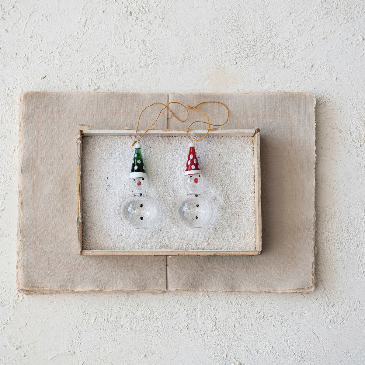 Set of two CREATIVE COOP HANDMADE GLASS SNOWMAN ORNAMENTS with gold strings—each features a round body, painted face with carrot nose, and a pointed hat (one red with white dots, one green).