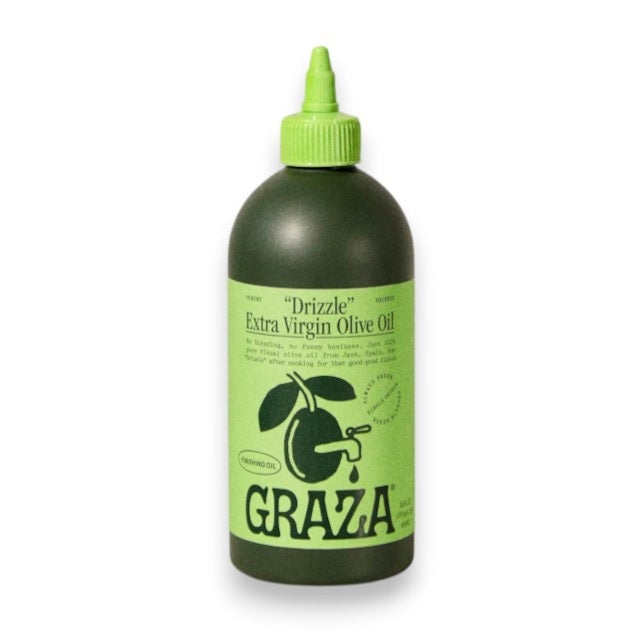A green squeeze bottle labeled “GRAZA - DRIZZLE OLIVE OIL” from GRAZA, a finishing oil made in Spain, stands upright on a light surface with a neutral textured background.
