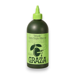 A green squeeze bottle labeled “GRAZA - DRIZZLE OLIVE OIL” from GRAZA, a finishing oil made in Spain, stands upright on a light surface with a neutral textured background.