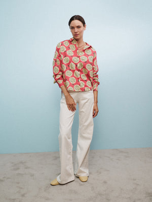 A woman sits on a clear chair with one arm propped on her head, wearing the 0039 ITALY LUCY BLOUSE IN A FRUIT PRINT, white pants, and yellow flats, against a light blue wall and gray carpet.