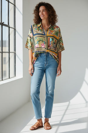 A person in a MOMONI - BROOKLYN blue shirt with a classic collar, paired with colorful beaded necklaces and tucked into striped pants. The loose-fitting, short-sleeve button-up stands out against the plain light background.