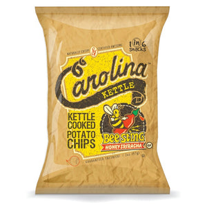 A 5-ounce bag of CAROLINA KETTLE - HONEY SRIRACHA CHIPS from 1 IN 6 SNACKS. The packaging features a vibrant design with a bee mascot holding a dripping honey stick and highlights the sweet and hot flavor, as well as emphasizing the natural crispiness and guaranteed freshness.