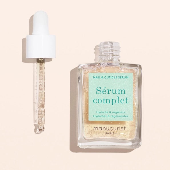 A square glass bottle labeled “MANUCURIST - COMPLETE SERUM” by MANUCURIST INC is displayed, featuring a dropper cap. The dropper, filled with a clear liquid dotted with tiny bubbles, rests beside the bottle—ideal for nourishing damaged nails and delivering essential hydration to cuticles. The scene is set against a soft beige background.