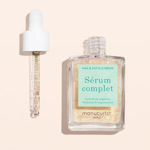 A square glass bottle labeled “MANUCURIST - COMPLETE SERUM” by MANUCURIST INC is displayed, featuring a dropper cap. The dropper, filled with a clear liquid dotted with tiny bubbles, rests beside the bottle—ideal for nourishing damaged nails and delivering essential hydration to cuticles. The scene is set against a soft beige background.