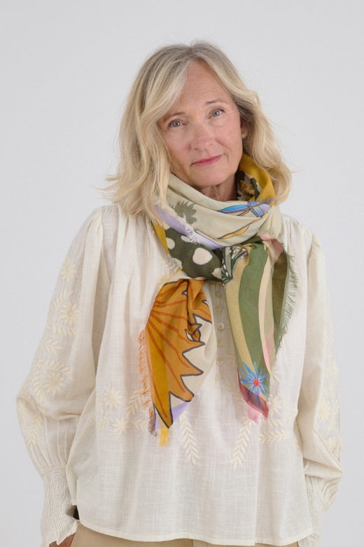 A woman holds the STORIATIPIC MALI MAX SCARF IN BEIGE, a luxurious cotton-modal accessory with colorful abstract and botanical patterns, against a plain white background.
