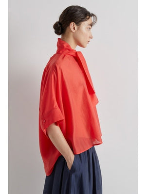 Wearing the MEIMEIJ Scarf Tie Neck Blouse in bright red, featuring a loose fit and wide short sleeves, paired with a dark pleated skirt, the person stands against a plain light background.